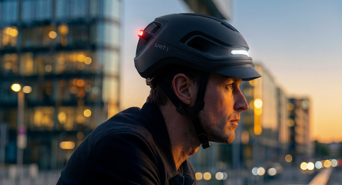 Radfahrer in der Dämmerung mit UNIT 1 AURA Helm und eingeschalteten Lichtern vor Stadtkulisse